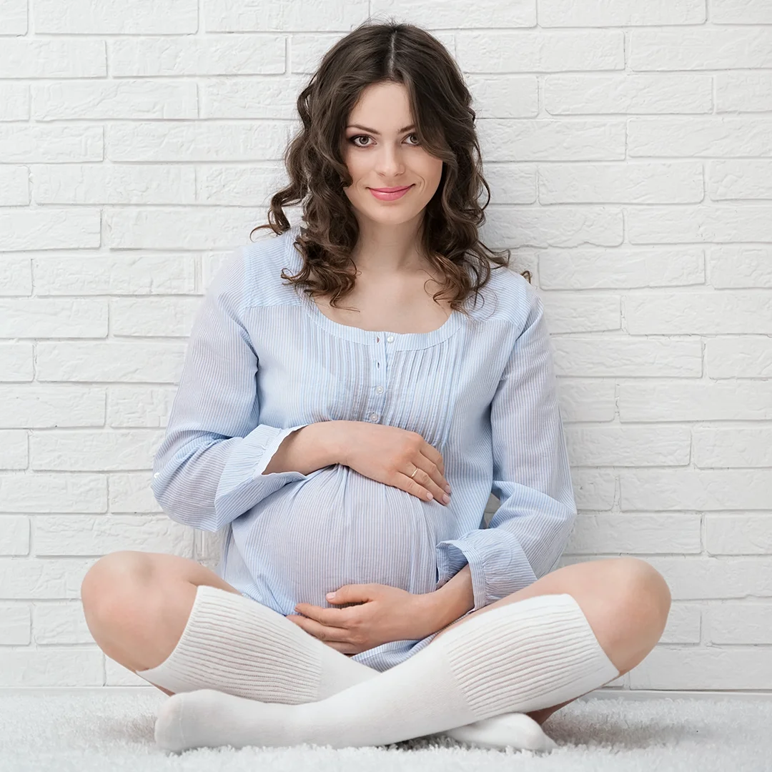 Uśmiechnięta kobieta w ciąży siedząca na podłodze w wygodnej pozycji, trzymająca ręce na brzuchu. Obrazek ilustruje tematykę flebologiczną związaną z ciążą, w której często dochodzi do zwiększonego ryzyka wystąpienia niewydolności żylnej oraz powstawania żylaków.